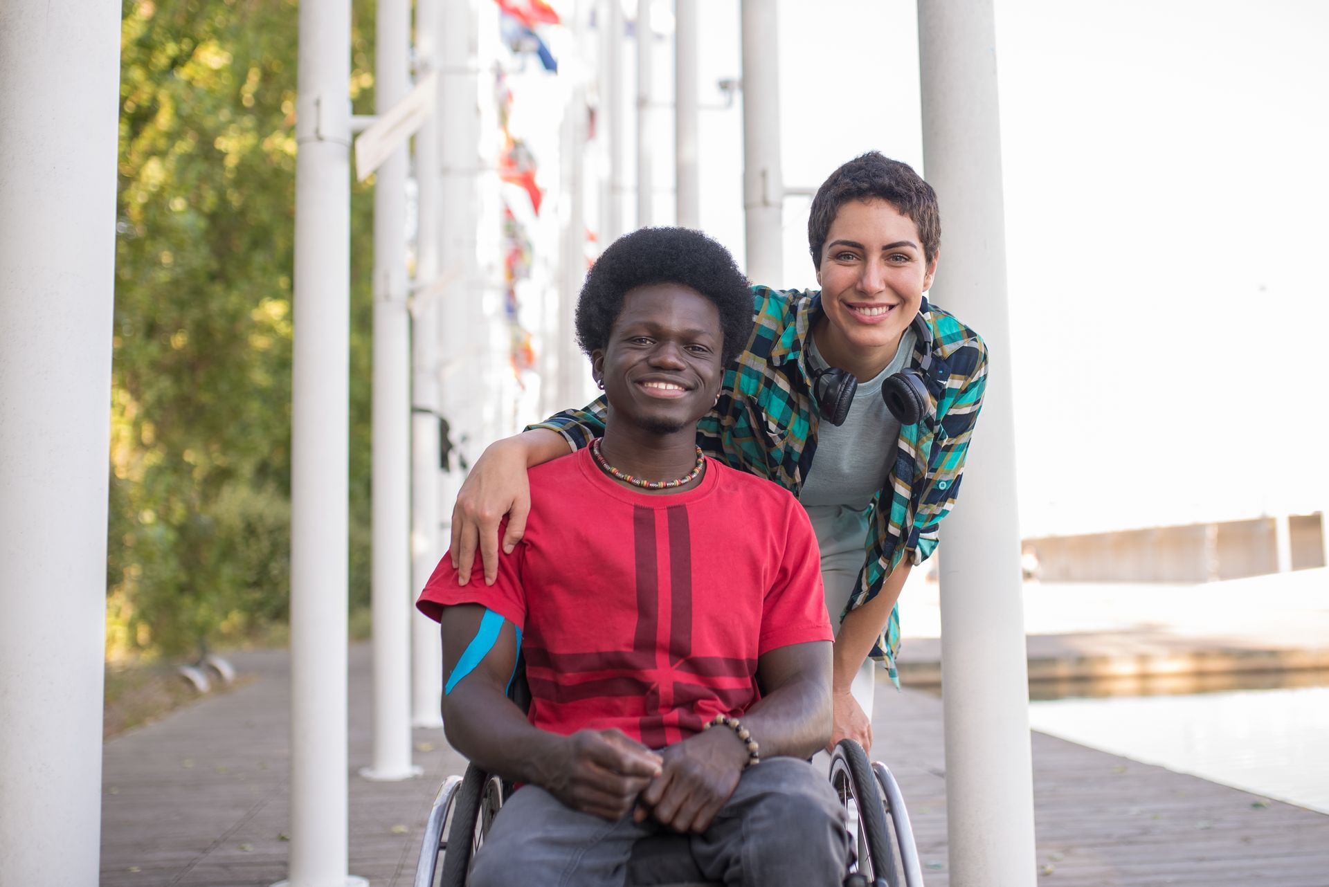 A smiling young man in a wheelchair with a friend leaning in beside him, her arm around his shoulder, outdoors on a sunny day